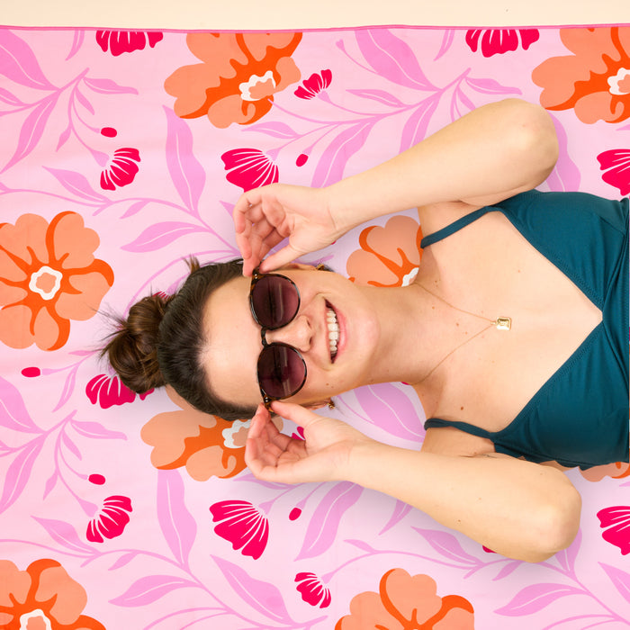A woman in sunglasses and a teal swimsuit smiles while lying on The Darling Effect’s Poppin' Petals Hot Pink XL Quick-Dry Beach Towel, holding her sunglasses with both hands.