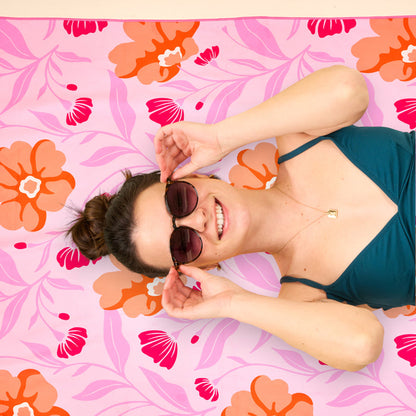 A woman in sunglasses and a teal swimsuit smiles while lying on The Darling Effect’s Poppin' Petals Hot Pink XL Quick-Dry Beach Towel, holding her sunglasses with both hands.