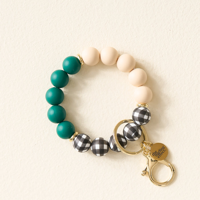 The Darling Effect Pretty in Plaid Hands-Free Keychain Wristlet features teal, cream, and black-and-white plaid silicone beads with gold key rings and a “she inspires others” charm, showcased on a light background.