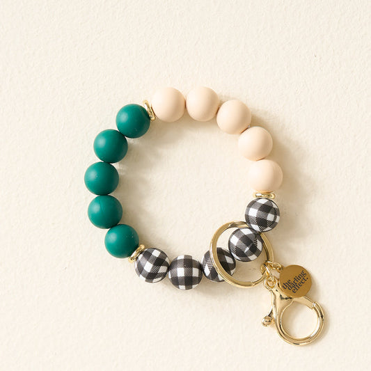 The Darling Effect Pretty in Plaid Hands-Free Keychain Wristlet features teal, cream, and black-and-white plaid silicone beads with gold key rings and a “she inspires others” charm, showcased on a light background.