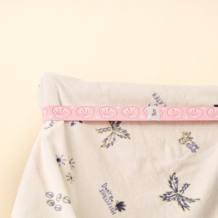A soft white fabric with blue and yellow floral patterns is neatly folded, wrapped at the top with a pink Put Towel Band-Seashells by the-darling-effect, featuring shell designs and a small white tag, set on a pale beige background.