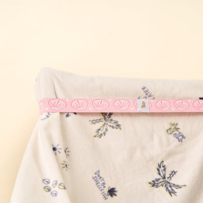A soft white fabric with blue and yellow floral patterns is neatly folded, wrapped at the top with a pink Put Towel Band-Seashells by the-darling-effect, featuring shell designs and a small white tag, set on a pale beige background.