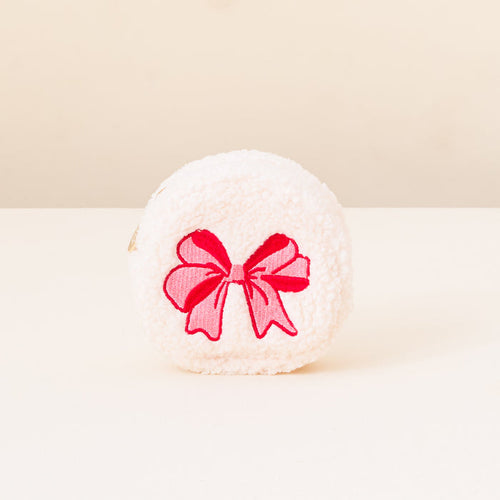 A round, fluffy white Red Bow Circle Teddy Pouch by The Darling Effect sits on a white surface with a light beige background, featuring a bright pink embroidered bow—a charming travel accessory for fans of Christmas teddy pouches.