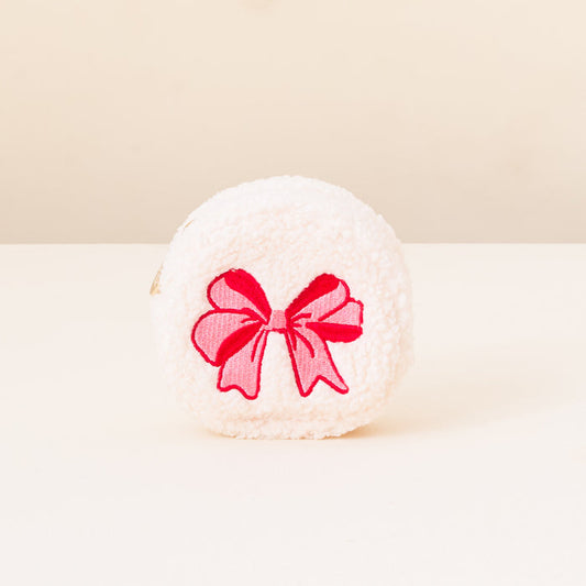 A round, fluffy white Red Bow Circle Teddy Pouch by The Darling Effect sits on a white surface with a light beige background, featuring a bright pink embroidered bow—a charming travel accessory for fans of Christmas teddy pouches.