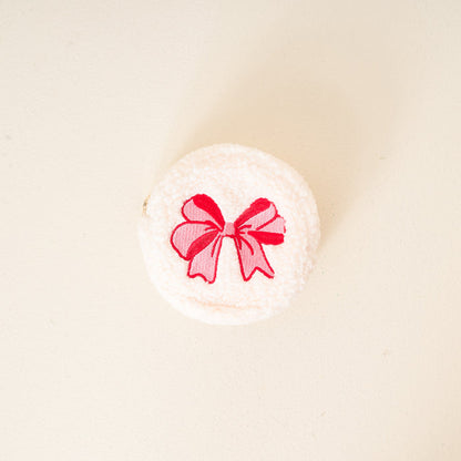 The Darling Effect's Red Bow Circle Teddy Pouch—a round, white, fluffy cosmetics case with an embroidered red bow—makes the perfect travel or festive accessory, shown against a simple light background.