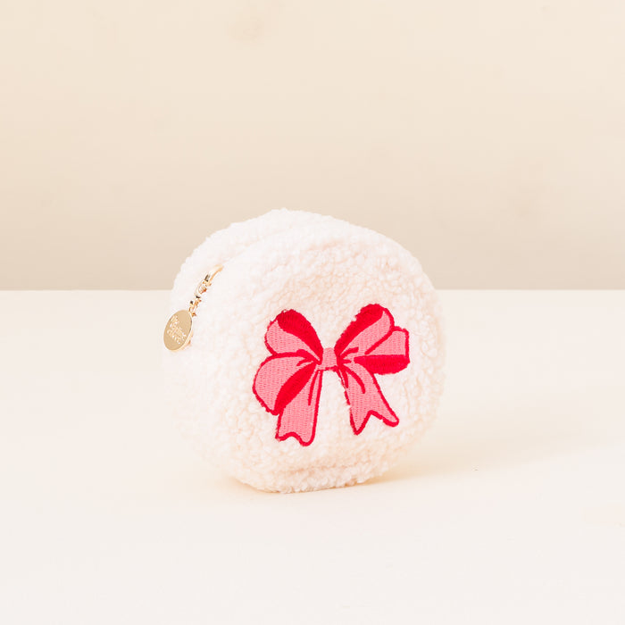The Darling Effect's Red Bow Circle Teddy Pouch—a round, white, fluffy pouch with pink and red bow design and a gold charm zipper—rests on a light surface against a cream background.