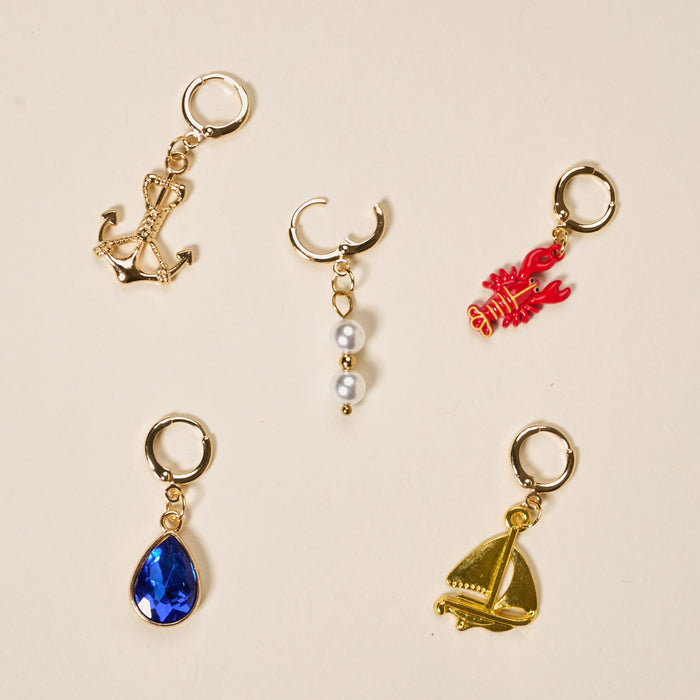 The Darling Effect’s Sail Away The Ultimate Charm Pack features five gold charms—a lobster, anchor, crescent moon with pearls, blue teardrop gem, and sailboat—each with an attachment loop. Ideal for charm collectors or DIY necklaces.