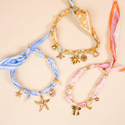 Three colorful statement necklaces from The Darling Effect’s Sail Away The Ultimate Charm Pack, featuring gold heart, bee, flower, starfish, seashell, and bow charms, are displayed on a cream background—perfect for playful accessorizing.