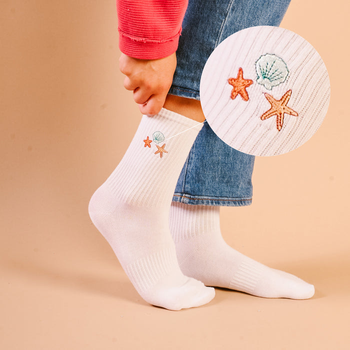 A close-up shows a person in blue jeans and a red rolled-sleeve top wearing The Darling Effect’s Seashells Embroidered Crew Socks, featuring colorful embroidered seashell and starfish designs.