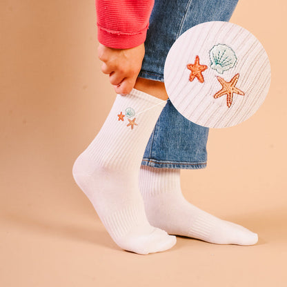 A close-up shows a person in blue jeans and a red rolled-sleeve top wearing The Darling Effect’s Seashells Embroidered Crew Socks, featuring colorful embroidered seashell and starfish designs.
