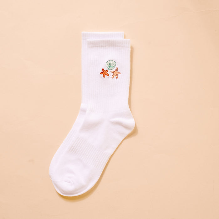 The Darling Effect Seashells Embroidered Crew Sock features a white women’s design with green seashell, orange starfish, and peach starfish embroidery near the top—perfect for fans of colorful socks. Displayed on a beige background.