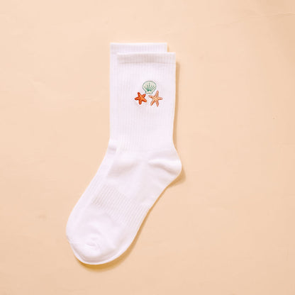 The Darling Effect Seashells Embroidered Crew Sock features a white women’s design with green seashell, orange starfish, and peach starfish embroidery near the top—perfect for fans of colorful socks. Displayed on a beige background.