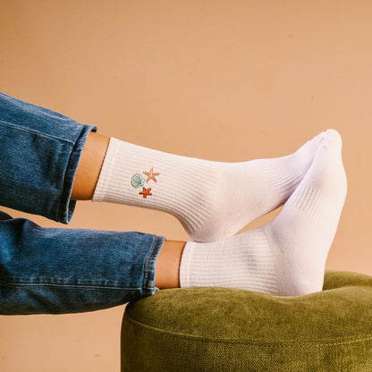 A person wears The Darling Effect’s Seashells Embroidered Crew Socks—white with small shell and starfish designs—paired with blue jeans while resting their feet on a green cushioned stool, all set against a beige background.