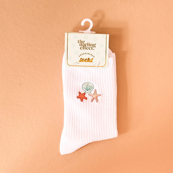 The Darling Effect’s Seashells Embroidered Crew Socks feature white ribbed fabric with colorful seashell and starfish details, displayed on a retail hook against a peach backdrop for a playful finishing touch.