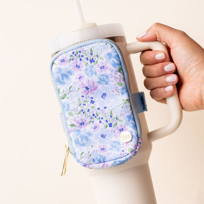 A hand with light pink nail polish holds a beige tumbler with a built-in straw, featuring the-darling-effect’s Secret Garden Blue Tumbler Fanny Pack—a pastel blue floral, water resistant accessory with a gold zipper.