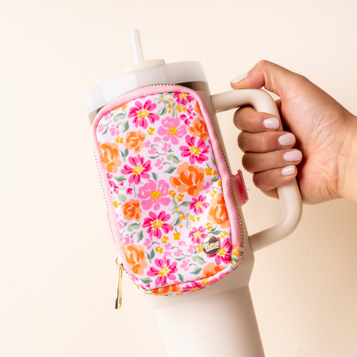 A hand holds a bestselling white tumbler with a straw, featuring the-darling-effect's Secret Garden Pink Tumbler Fanny Pack in pink floral print with pink, orange, and yellow flowers and gold zipper hardware attached to the front.