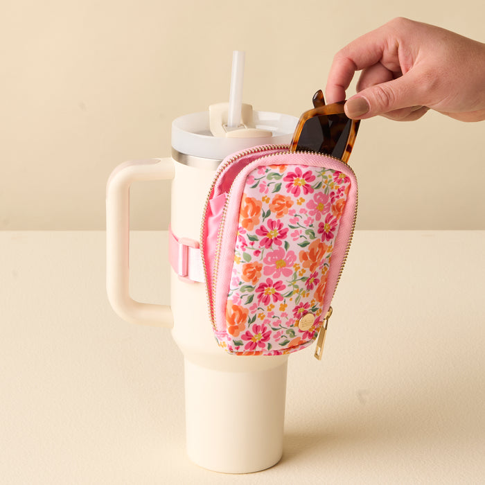 A hand places sunglasses into the-darling-effect's Secret Garden Pink Tumbler Fanny Pack, a floral-patterned, water-resistant pouch attached to a cream tumbler with a pink strap.