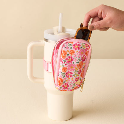 A hand places sunglasses into the-darling-effect's Secret Garden Pink Tumbler Fanny Pack, a floral-patterned, water-resistant pouch attached to a cream tumbler with a pink strap.