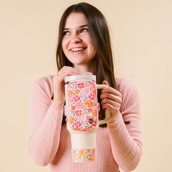 A smiling woman in a pink ribbed sweater holds the-darling-effect's Secret Garden Pink Tumbler, featuring a vibrant floral design and a protective silicone boot, against a beige background.