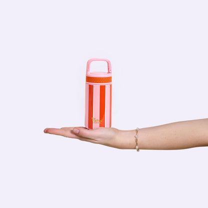 A hand wearing a bracelet holds The Darling Effect’s Sicily Stripes Pink 6 oz Lil' Bitty Bottle—red and white striped with a pink lid and handle—against a plain light background, ideal for hydration on the go.
