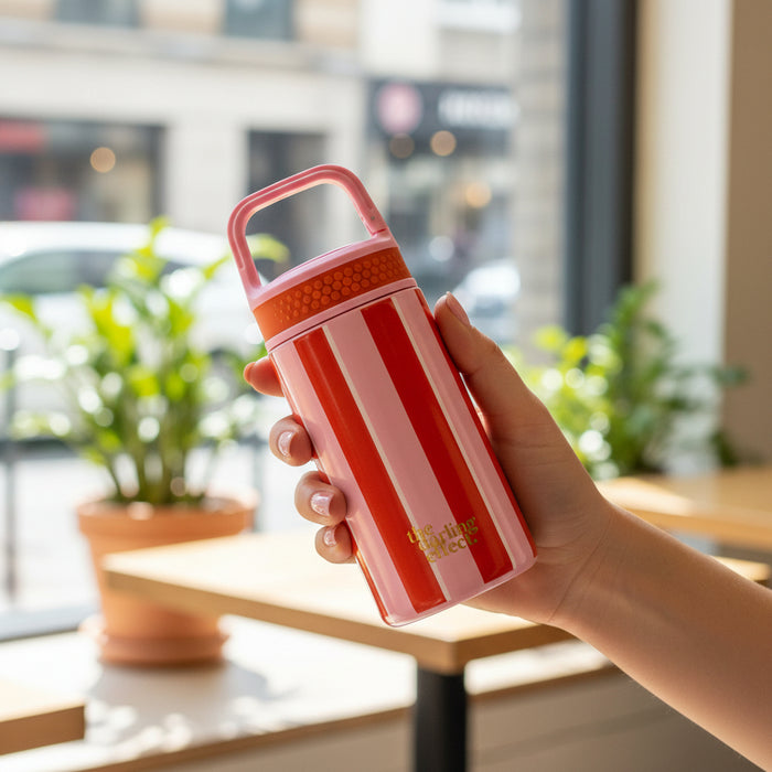 A hand holds The Darling Effect's Sicily Stripes Pink 6 oz Lil' Bitty Bottle—its red and white stripes and handle make it perfect for hydration on the go, shown indoors by a window with potted plants and blurred outdoor scenery.