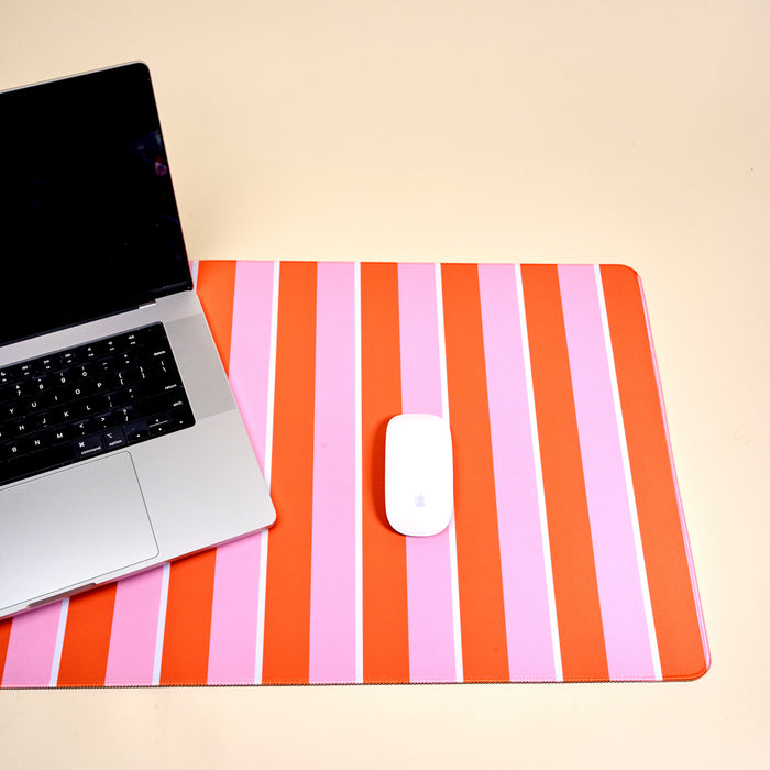 A silver laptop and a white wireless mouse rest on The Darling Effect's Sicily Stripes Pink Style Station Desk Mat, featuring vertical red and pink stripes, atop a light beige surface.