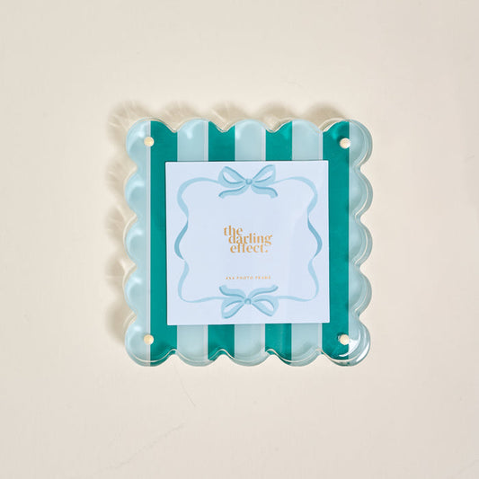 The Sicily Stripes Teal Square Scalloped Acrylic Picture Frame by The Darling Effect features teal and light blue striped scalloped edges and a magnetized design. The center card displays "the darling effect" in gold with blue bow illustrations.