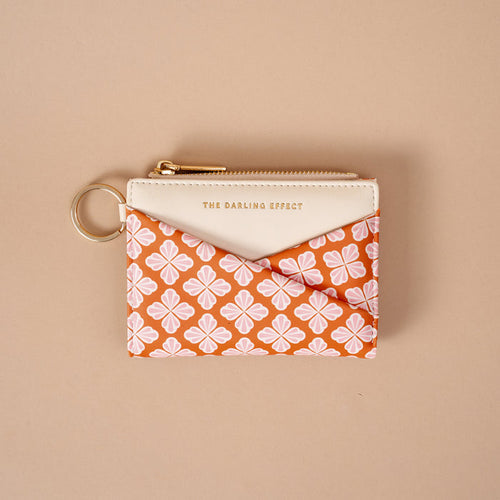 Introducing the Sienna Spice Luxe Pocket Wallet by The Darling Effect—a compact zippered wallet with a key ring, designed with a cream top and an orange base decorated in a white and pink floral pattern. Crafted from vegan leather for both style and sustainability, this pouch features “THE DARLING EFFECT” embossed in gold on the cream section, offering an elegant finishing touch.