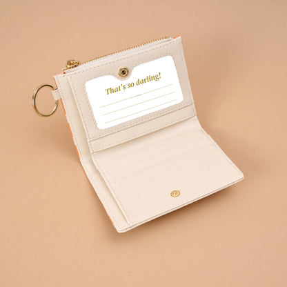 The Darling Effect Sienna Spice Luxe Pocket Wallet, crafted from cream-colored vegan leather and featuring a keyring, is shown open to reveal a card slot with a card that reads “That’s so darling!” against a beige backdrop, highlighted by an elegant criss-cross design.
