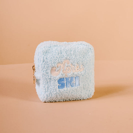 A light blue, fuzzy Square Aprés Ski Teddy Pouch by The Darling Effect rests on a beige surface. “Après SKI” is embroidered on the front in white and blue letters, making this cozy pouch perfect for storing your après-ski essentials.