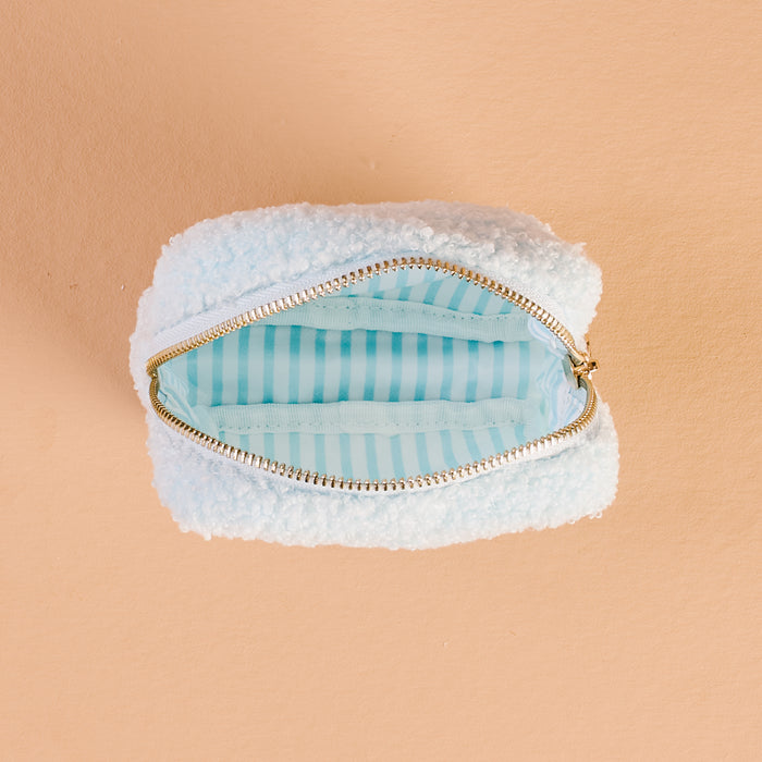 The Darling Effect’s Square Aprés Ski Teddy Pouch features a small, fluffy white sherpa exterior and a gleaming gold zipper, opening to reveal a light blue interior with crisp white stripes. Set against a peach-colored background, it’s the perfect accessory for stashing your essentials après-ski.