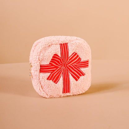 The Darling Effect’s Square Holiday Gift Bow Red Teddy Pouch is a light pink, fuzzy sherpa pouch with a large red bow and side zipper—perfect as a festive organizer for the holidays. Photographed on a plain beige background.