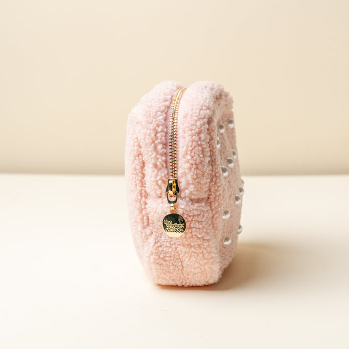 The Darling Effect’s Square Pearl Blush Teddy Pouch, a small pink sherpa pouch with silver studs and a gold zip, stands upright on a cream surface against a pale background.