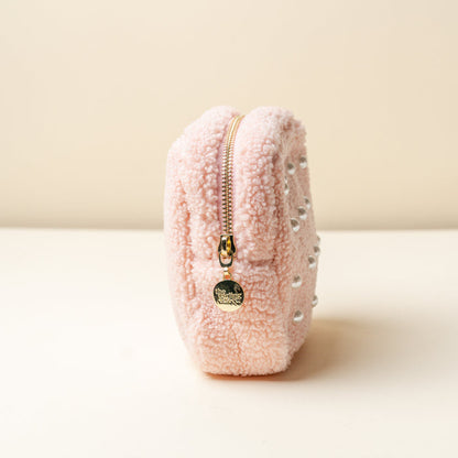 The Darling Effect’s Square Pearl Blush Teddy Pouch, a small pink sherpa pouch with silver studs and a gold zip, stands upright on a cream surface against a pale background.