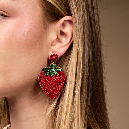 Close-up of a woman with straight blonde hair tucked behind her ear, showcasing The Darling Effect’s Strawberry Beaded Earrings—large, hand-beaded accessories featuring red and green details—against a plain beige background.