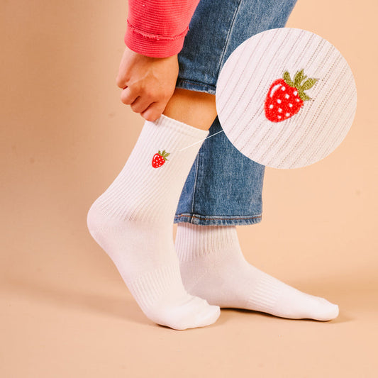 A close-up shows a person wearing The Darling Effect’s Strawberry Embroidered Crew Socks, featuring tiny strawberries. The colorful socks are styled with rolled-up jeans and a pink long-sleeve shirt.