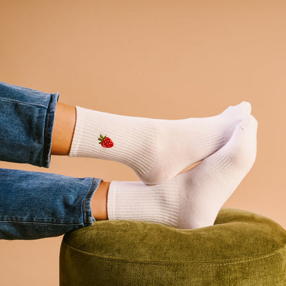 The Darling Effect's Strawberry Embroidered Crew Socks feature a tiny strawberry on the ankle. One colorful sock is paired with blue denim jeans, while the other rests on a green ottoman.