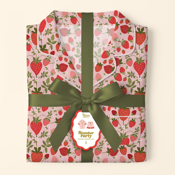 Folded pink Strawberry Fields Slumber Party Pajama Shorts Set by The Darling Effect, featuring a red strawberry and green leaf pattern on ultra-soft fabric. Tied with an olive green ribbon, the tag shows a pajama illustration.
