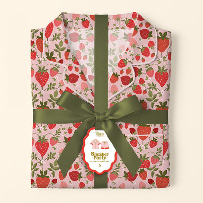 Folded pink Strawberry Fields Slumber Party Pajama Shorts Set by The Darling Effect, featuring a red strawberry and green leaf pattern on ultra-soft fabric. Tied with an olive green ribbon, the tag shows a pajama illustration.
