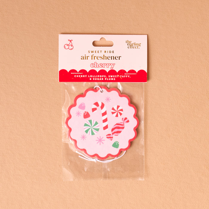 The Darling Effect’s Sugar Rush Sweet Ride Car Air Freshener features a round, scalloped edge design with candy canes, lollipops, and sweets on pink. Packaged in clear plastic, it fills your car with a long-lasting cherry scent.