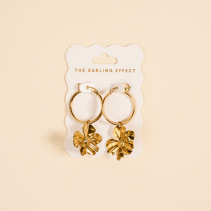 Sunlit Petal Standout Earrings by The Darling Effect feature high-shine golden finish and dangling flower charms, displayed on a white card—perfect for special occasions against a soft beige background.