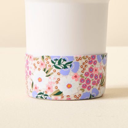 A white cup with a colorful floral base sits on a light surface, protected by The Darling Effect Sweet Meadow Purple Tumbler Silicone Boot for added style and stability.