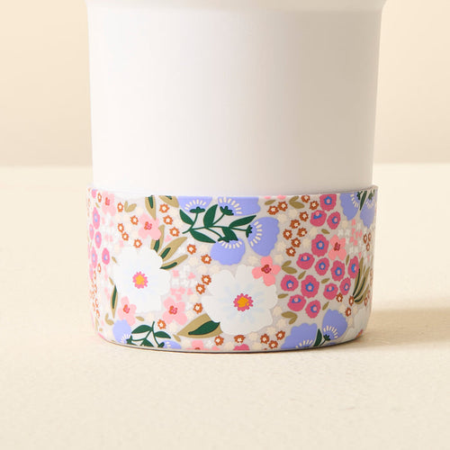 A white cup with a colorful floral base sits on a light surface, protected by The Darling Effect Sweet Meadow Purple Tumbler Silicone Boot for added style and stability.