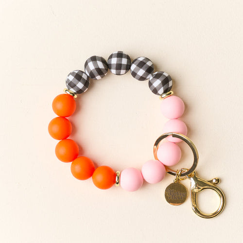 The Darling Effect’s Sweet & Spooky Hands-Free Keychain Wristlet features orange, pink, and black-and-white checkered silicone beads with a gold keyring and charm—ideal for keeping keys stylishly accessible on your wrist.
