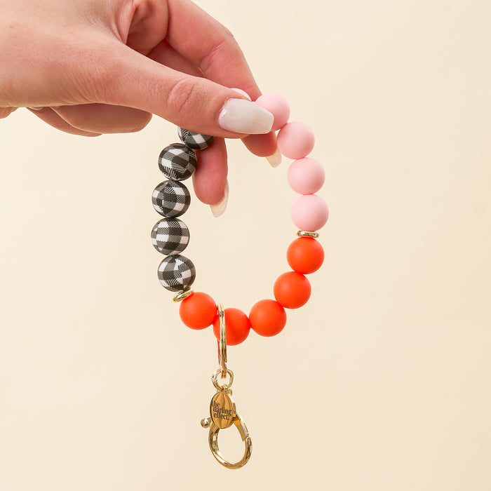 A manicured hand holds The Darling Effect’s Sweet & Spooky Hands-Free Keychain Wristlet in black plaid, pink, and orange with a gold clasp and engraved tag, standing out against a plain light beige background.