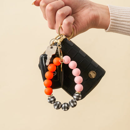 A hand holds The Darling Effect's Sweet & Spooky Hands-Free Keychain Wristlet, featuring two silver keys, a small black checkered wallet, and a keychain with orange, pink, and black-and-white silicone beads.