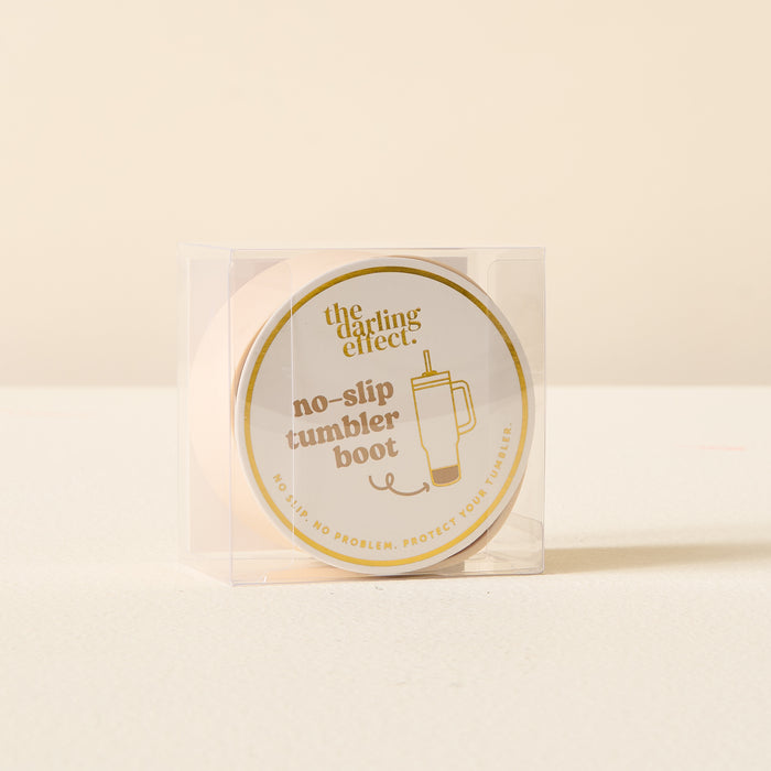 A clear plastic box holds The Darling Effect's Tan Tumbler Silicone Boot, a round, beige no-slip boot for tumbler protection. The cream label has a gold logo and simple tumbler illustration.