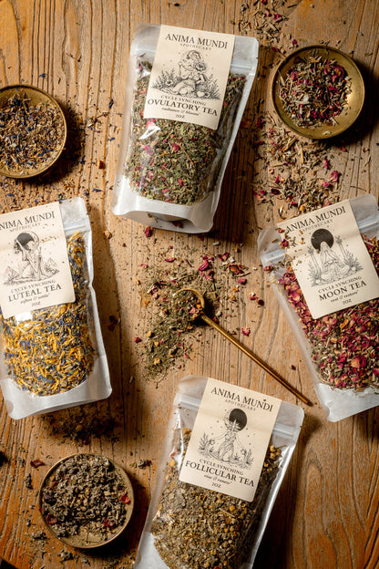Four stand-up pouches of Anima Mundi herbal teas (Ovulatory Tea, Luteal Tea, Pollinator Tea, Moon Tea) arranged on a rustic wooden table with small bowls of loose herbal ingredients and a wooden spoon.