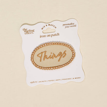 The Darling Effect’s Things Mix & Match Iron-On Patch features an oval shape with gold trim and “Things” in gold script, displayed on a branded card—ideal for adding a stylish, colorful touch to your jackets.