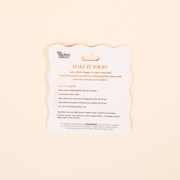 A square instruction card with scalloped edges and orange text, titled MAKE IT YOURS, guides you on using The Darling Effect’s Things Mix & Match Iron-On Patch to customize jackets. Includes steps, a warning, and website info at the bottom.